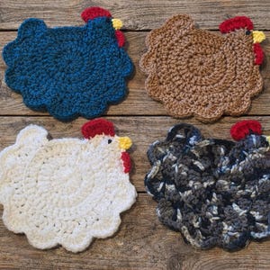 May include: Four handmade crochet chicken-shaped trivets in various colors: blue, brown, white, and a mix of gray and black. Each trivet features a red comb and a yellow beak. The trivets are placed on a rustic wooden surface.