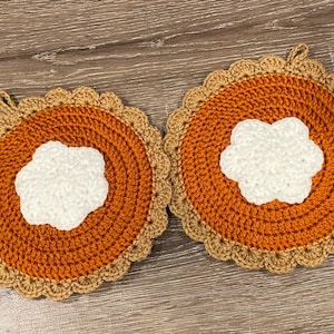 Peut inclure: Deux dessous de plat faits à la main au crochet en forme de parts de tarte à la citrouille. Le centre est orange, avec un motif de crème fouettée blanche. La croûte est beige et chacune a une boucle pour suspendre. Un accessoire de cuisine douillet.