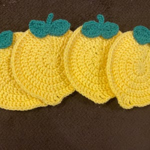 May include: Four crocheted lemon-shaped coasters with green leaves. The coasters are yellow and green.