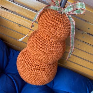 May include: An orange, crocheted pumpkin decoration with a green and red plaid ribbon bow. The pumpkin has a stacked design and a dark metal stem. The pumpkin is resting on a blue cushion.
