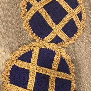 May include: Two handmade crochet coasters. Each coaster features a navy blue center with a tan lattice design and scalloped edge, resembling miniature pies. The coasters are placed on a wooden surface.