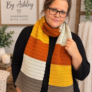 May include: A long, rectangular crochet scarf featuring horizontal color-blocked stripes in mustard yellow, burnt orange, cream, and dark gray. The scarf is crafted with a consistent textured stitch pattern and is draped around the neck.