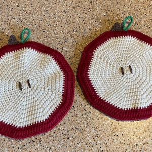 May include: Two handmade crochet apple-shaped pot holders. Each has a white center, a red border, and brown and green accents to resemble an apple. The pot holders are designed to protect surfaces from heat.