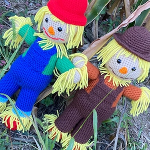 May include: Two handmade crochet scarecrows. One wears a red hat and blue overalls, the other a brown hat and overalls. Both have yellow straw hair, orange noses, and black eyes. They are lying in a grassy area.