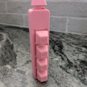 May include: A vertical pink plastic device featuring four protruding square buttons arranged in a column, each marked with the numbers 2, 3, 4, and 5. The structure has a rectangular body with a small, separate top piece connected by a thin bridge.