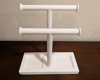 May include: A white, 3D-printed jewelry display stand. The stand has a rectangular base, a central vertical support, and two horizontal bars for hanging necklaces or bracelets. The stand is made of white plastic and sits on a dark brown wooden surface.