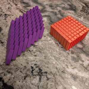 May include: Two 3D-printed geometric shapes. One is a purple, angled rectangular prism. The other is a red-orange cube. Both are composed of many small, rectangular blocks. The objects are on a speckled, gray surface.