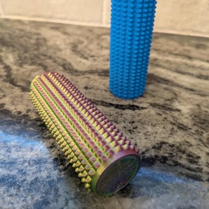 Peut inclure: Deux rouleaux de massage cylindriques avec une surface texturée et pointue. L'un des rouleaux est d'un bleu vif uni, tandis que l'autre présente une finition métallique irisée qui varie entre des tons de vert, d'or et de violet.