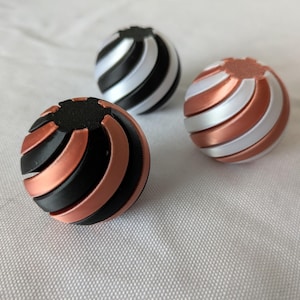 Puede incluir: Tres objetos esféricos con un diseño en espiral. Uno es negro y cobrizo, otro es negro y plateado, y el tercero es cobrizo y plateado. Los objetos están sobre una superficie blanca.
