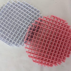 May include: Two circular, grid-like objects. One is white, the other is pink. The objects are made of a series of intersecting lines, creating a square pattern. The objects are likely made of plastic.