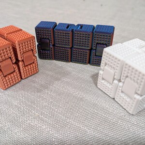 May include: Three fidget cubes in orange, blue, and white. The orange cubes are side by side, the blue cubes are stacked, and the white cube is in the foreground. Each cube has a textured surface.