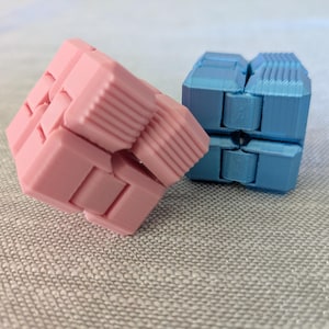 May include: Two fidget cubes, one pink and one blue, are displayed. The pink cube is partially unfolded, revealing its segmented design. The blue cube is fully formed. Both cubes have a textured, ridged surface on some of their segments.