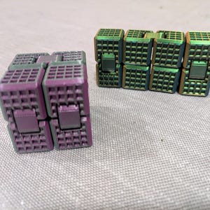 May include: Two fidget cubes with a grid-like pattern. One cube is purple and the other is green and gold. The cubes are made of metal and have a square shape. The cubes are on a gray surface.
