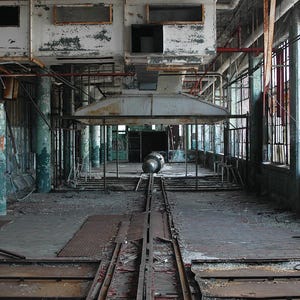 May include: A view of an abandoned factory with exposed brick walls, rusted metal beams, and a large metal structure in the center. The factory floor is covered in dirt and debris, and there are several sets of rusty railroad tracks leading to the metal structure.