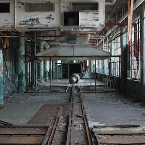 May include: A view of an abandoned industrial building with exposed brick walls, rusted metal beams, and a large metal structure in the center. The building has large windows with metal bars and a concrete floor with a set of tracks running down the center.