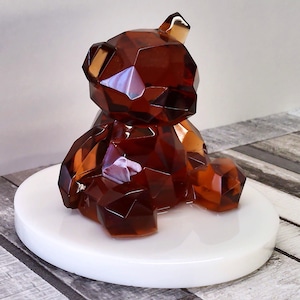 Può includere: Una statuetta a forma di orso sfaccettato, color ambra, su una base circolare bianca. Il design geometrico dell'orso crea un effetto scintillante, con la luce che si riflette sulle sue molteplici superfici. Un oggetto decorativo moderno.