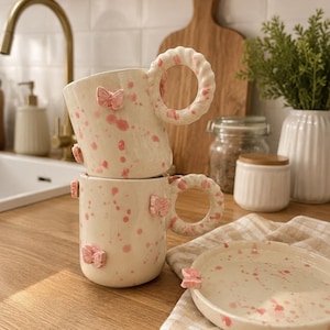 Pode incluir: Duas canecas de cerâmica empilhadas verticalmente e um pires a condizer, todas com uma base de cor creme com salpicos de tinta rosa e pequenos relevos de borboletas cor-de-rosa. Cada caneca tem uma pega circular espessa com bordo ondulado.