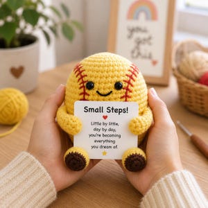 Könnte beinhalten: Ein gelber gehäkelter Baseball mit roten Nähten, schwarzen Augen und einem lächelnden Mund. Er hält eine weiße Karte mit dem Text "Small Steps!" und einer inspirierenden Botschaft. Der Baseball hat kleine, braune Füße.