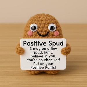 May include: A crocheted brown potato-shaped figure with large googly eyes and pink cheeks holds a white sign. The sign reads "Positive Spud" and includes an inspirational message. The figure has small arms and a smiling mouth.