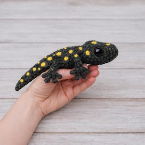 Peut inclure: Un lézard en crochet fait à la main, de couleur gris foncé avec des taches jaunes. Le reptile en peluche a des yeux noirs et de petites pattes. Le jouet est tenu dans une main sur un fond en bois clair. L'objet est unique et fait à la main.