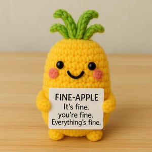 May include: A crocheted yellow pineapple with a green leafy top, black eyes, and pink cheeks. The pineapple holds a white sign that reads "FINE-APPLE It's fine. you're fine. Everything's fine."