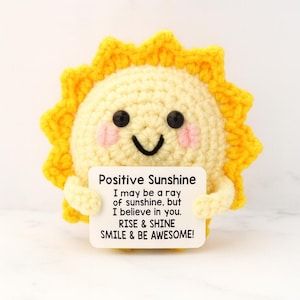 May include: A crocheted sun with a smiling face, rosy cheeks, and yellow rays. The sun holds a sign that reads "Positive Sunshine" with an inspirational message. A cheerful, handmade item.