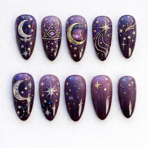 May include: A set of ten almond-shaped, dark purple press-on nails with celestial designs. Each nail features gold and silver accents, including moons, stars, and constellations. Some nails have glitter and a gradient effect.