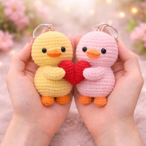 Puede incluir: Dos llaveros de pato de ganchillo hechos a mano, uno amarillo y otro rosa, cada uno sosteniendo un corazón rojo. Los patos tienen picos y patas naranjas y ojos negros. Los llaveros se sostienen en las manos de una persona, con un fondo borroso de flores rosas.