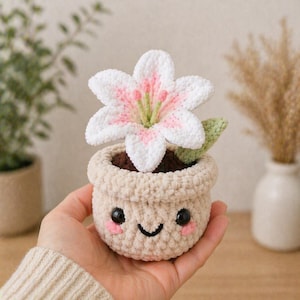 May include: A crocheted flower in a pot. The white flower has pink accents and green stamens. The pot is beige with a smiling face design. The flower pot is a handmade item.