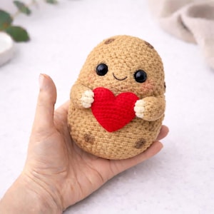 May include: A crocheted potato-shaped toy with a smiling face, black eyes, and a red heart. The toy is tan with brown spots and small arms with white tips. The toy is held in a hand.