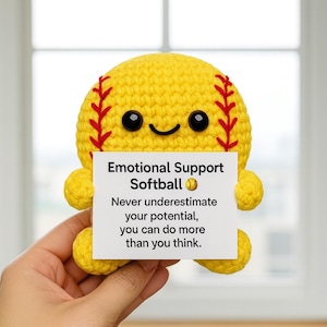 Könnte beinhalten: Ein gelber gehäkelter Softball mit roten Nähten und schwarzen Augen, der ein weißes Schild hält. Auf dem Schild steht "Emotional Support Softball" und enthält eine inspirierende Botschaft. Der Softball hat kleine gelbe Anhänge.