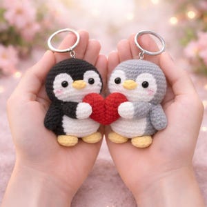 Puede incluir: Dos llaveros de pingüinos hechos a mano con ganchillo, uno en blanco y negro, el otro en gris y blanco, cada uno sosteniendo un corazón rojo. Los pingüinos tienen patas amarillas y se sostienen en manos abiertas. Los llaveros tienen anillos plateados.