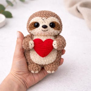 May include: A handmade crochet sloth doll in shades of brown, cream, and red. The sloth is holding a red heart. The doll has large black eyes and a smiling face. The sloth is being held in a person's hand.