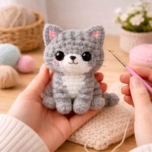 May include: A handmade crochet cat in gray and white, with pink accents on the ears and cheeks. The cat has large black eyes and a small, smiling mouth. The cat is sitting on a crocheted surface.