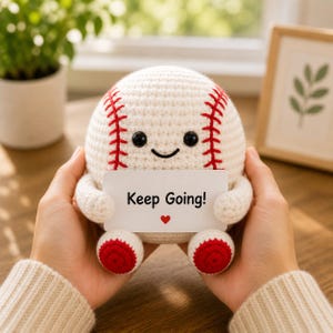 Könnte beinhalten: Ein gehäkelter Baseball mit einem lächelnden Gesicht und roten Nähten. Der Baseball hält eine weiße Karte mit dem Text "Keep Going!" und einem roten Herzen. Der Baseball hat rote Akzente an den Füßen.