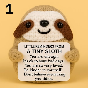 May include: A crocheted sloth figurine with a white sign. The sloth is tan and white with brown accents. The sign reads "LITTLE REMINDERS FROM A TINY SLOTH" followed by positive affirmations. A number 1 is in the top left corner.