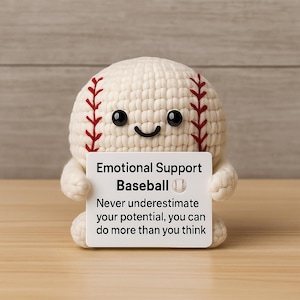 Könnte beinhalten: Ein gehäkelter Baseball mit einem lächelnden Gesicht und schwarzen Augen, der ein Schild mit der Aufschrift "Emotional Support Baseball" hält. Das Schild enthält auch den Text "Never underestimate your potential, you can do more than you think."