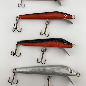 Könnte beinhalten: Vier Vintage-Angelköder in einer Reihe. Der obere Köder ist rot und weiß, der zweite ist rot, der dritte ist schwarz und rot und der untere Köder ist silberfarben. Jeder Köder hat zwei Drillingshaken und eine Metallflosse.