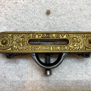 Puede incluir: Nivel Stanley vintage con carcasa de metal dorado ornamentada. El nivel tiene un indicador de burbuja y el texto "STANLEY PAT JUNE 23. 96." Está montado sobre una base de metal oscuro con un tornillo de ajuste.