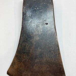 May include: Aged axe head with a dark, worn look. The metal is dark grey with rust and discolouration, indicating age and use. The axe head features a broad, curved blade and a rectangular top. The axe head is approximately 20 cm long.