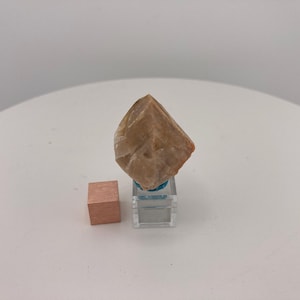 Puede incluir: Un cristal marrón claro con forma de pirámide, exhibido sobre un soporte de acrílico transparente. Un pequeño cubo color cobre está a la izquierda. Los objetos están sobre una superficie blanca.