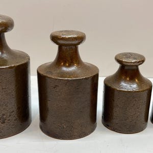 Puede incluir: Un conjunto de cuatro pesas cilíndricas de metal de tamaños graduados con una pátina marrón oscura y desgastada. Cada pesa presenta una base plana, un cuerpo vertical y un asa redondeada en la parte superior, dispuestas de mayor a menor.
