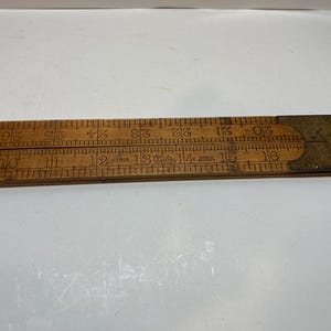 Pode incluir: Uma régua dobrável de madeira vintage com marcações numéricas pretas de 10 a 26. A ferramenta inclui um mecanismo de dobradiça metálica no lado direito com o texto "Elite" e "Made in Germany" claramente visível na superfície de madeira.