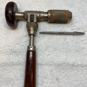 Puede incluir: Un taladro manual antiguo con un mango de madera oscura y un cuerpo de metal. El taladro tiene una perilla circular de madera y un agarre texturizado. Un destornillador de cabeza plana se coloca junto al taladro.