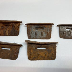 Puede incluir: Cinco objetos metálicos rectangulares desgastados con una pátina marrón óxido oscuro. Cada pieza tiene una pequeña abertura rectangular y una parte superior ligeramente saliente. Los objetos parecen envejecidos y posiblemente antiguos.