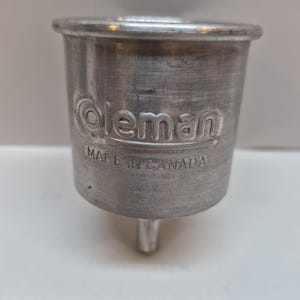 May include: A silver-colored metal funnel with a cylindrical body and a tapered spout at the bottom. The exterior features the embossed text Coleman and the words MADE IN CANADA positioned directly underneath the brand name in a clear font.