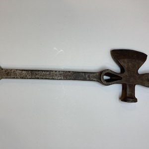 Könnte beinhalten: Ein Vintage-Multitool mit Hammer, Axt und Nagelzieher. Das Werkzeug besteht aus gealtertem Metall mit dunklem, rostigem Finish. Es hat einen langen Griff mit einem Loch am Ende zum Aufhängen.