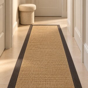 Peut inclure: Un tapis de couloir rectangulaire composé d'un centre en fibres naturelles tissées de couleur beige et d'une large bordure en tissu gris anthracite foncé. Le tapis est disposé en longueur sur un sol clair, révélant un tissage texturé.