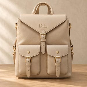 May include: A beige leather backpack featuring a top handle, a fold-over flap with a central buckle closure, and two front pockets with buckle straps. The letters D.L are embossed on the flap. The bag includes gold-toned metal hardware throughout.