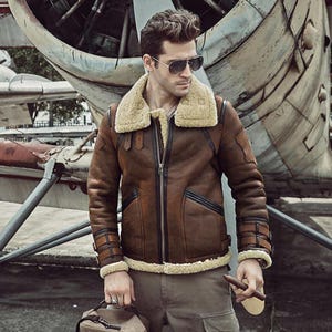 Puede incluir: Chaqueta de cuero marrón con cuello y forro de borrego. La chaqueta tiene cremallera, múltiples bolsillos y detalles de hebillas en las mangas. La chaqueta se usa con gafas de sol y pantalones color caqui.